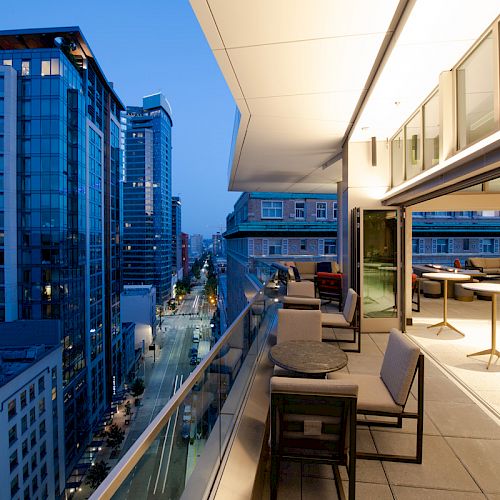 A modern cityscape is seen from a high-rise balcony featuring outdoor seating and tables, overlooking the street and other buildings at dusk.