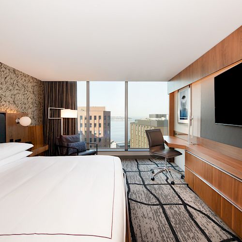 A modern hotel room features a large bed, a desk with a chair, a large TV, and a window with a city view and water in the distance.