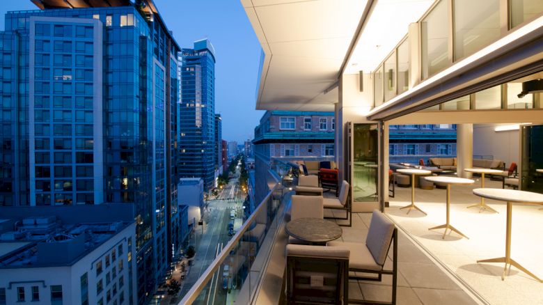 The image shows a modern balcony with chairs and tables overlooking a city street lined with tall buildings at dusk.