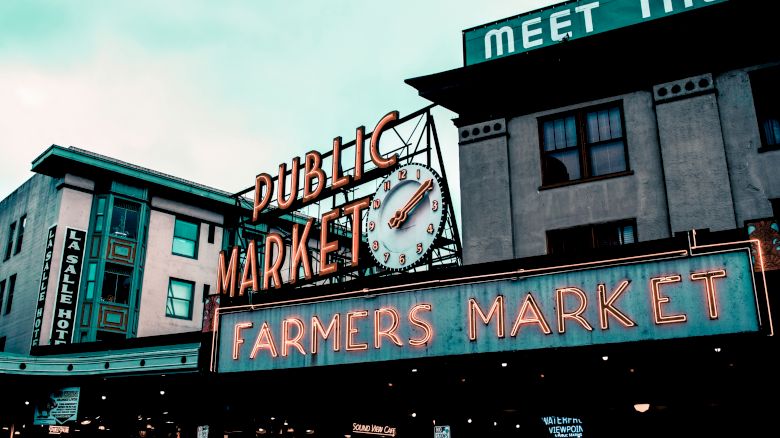 The image shows an iconic neon sign for a "Public Market" and "Farmers Market," set against older buildings with urban architecture.