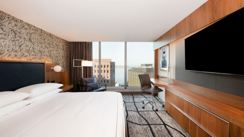 A modern hotel room with a large bed, flat-screen TV, desk, and window offering a city view. The room features sleek wood and contemporary decor.