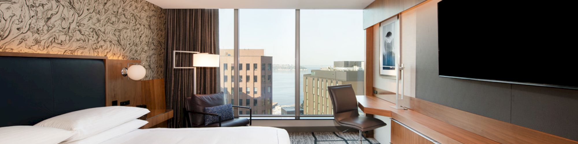 A modern hotel room with a large bed, desk, chair, wall-mounted TV, floor-to-ceiling window, and a view of city buildings and water in the distance.