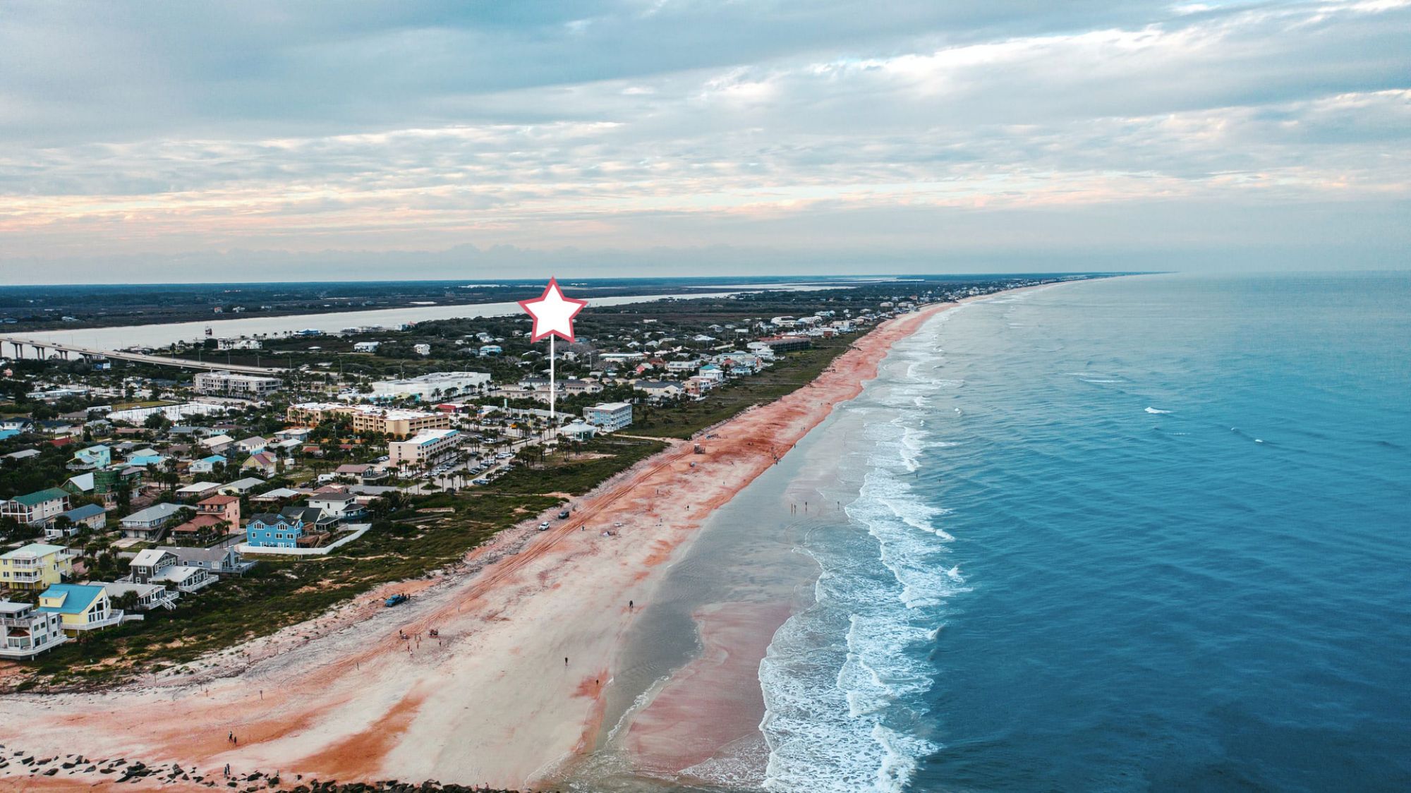 An aerial view of a coastline with a sandy beach, nearby buildings, and a star marker indicating a specific location on the shore.