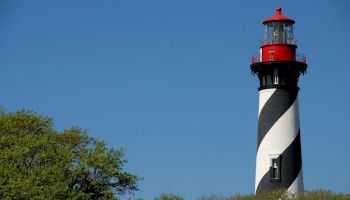 The image shows a black and white striped lighthouse with a red top, surrounded by green foliage under a clear blue sky.