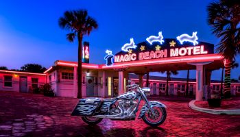 An illuminated "Magic Beach Motel" sign, with neon dolphins and stars, stands over a vintage-style building and a parked motorcycle.