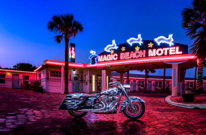 An illuminated "Magic Beach Motel" sign, with neon dolphins and stars, stands over a vintage-style building and a parked motorcycle.