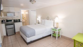 A neatly arranged bedroom includes a bed, side tables, lamps, a small kitchenette with appliances, and a green laundry basket on tiled flooring.