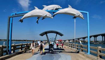 People walking on a pier under an arch with three dolphin sculptures, near water and a bridge in the background.