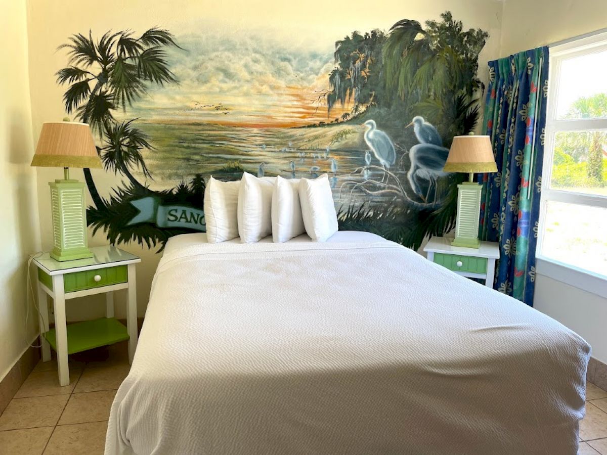 A cozy bedroom with a mural of a tropical sunset and birds, a bed with white linens and pillows, green nightstands with lamps, and a window.