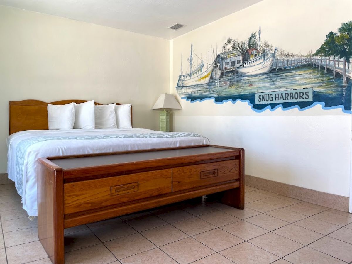 The image shows a tidy hotel room featuring a bed with four pillows, a wooden bench at the foot, a lamp, and a mural of Snug Harbor boats.