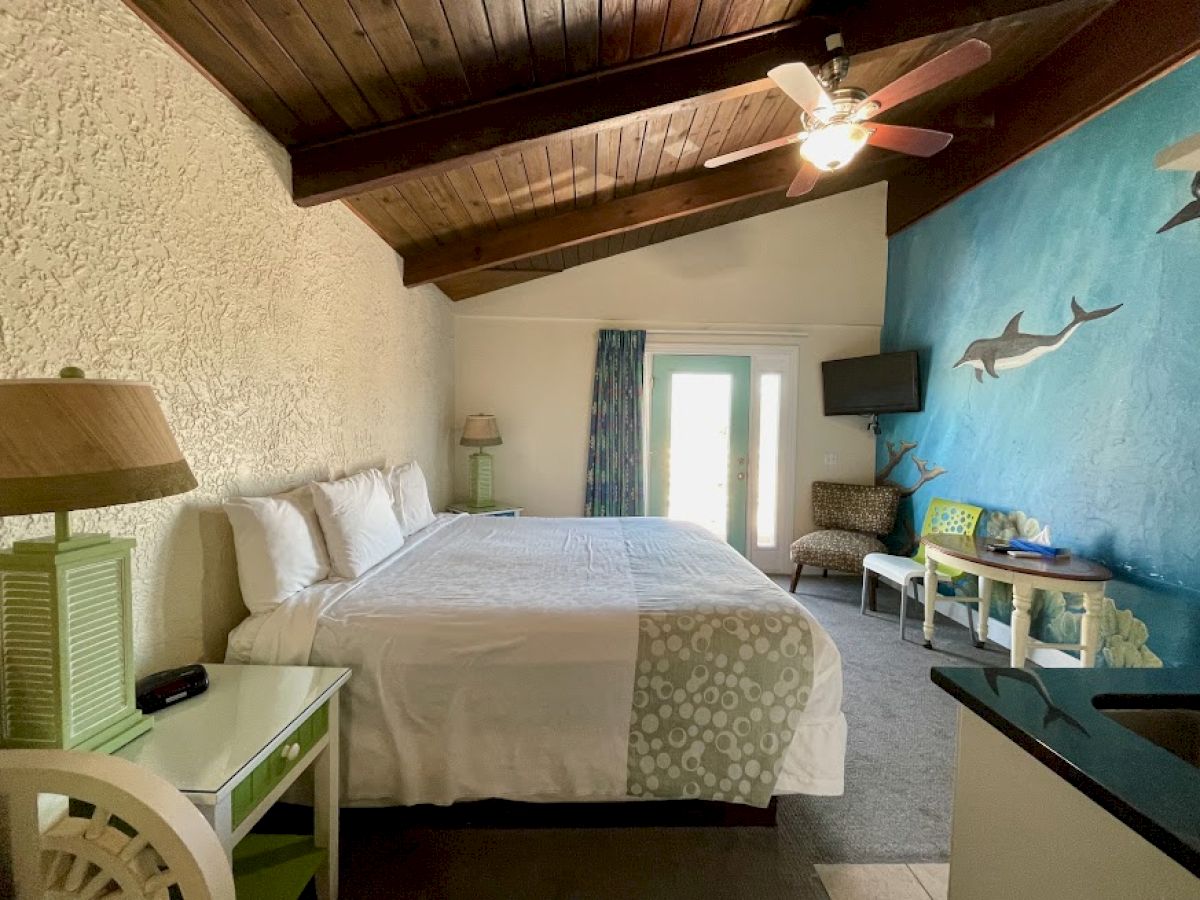 The image shows a cozy bedroom with a large bed, wooden ceiling, blue accent wall with fish decorations, a TV, and a small dining table.