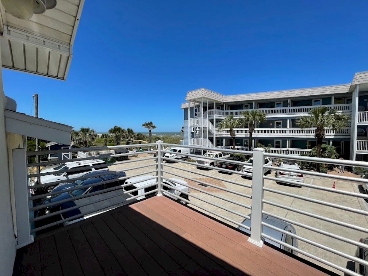 View from a balcony overlooking a parking lot, with a building complex and palm trees in the background under a clear blue sky.