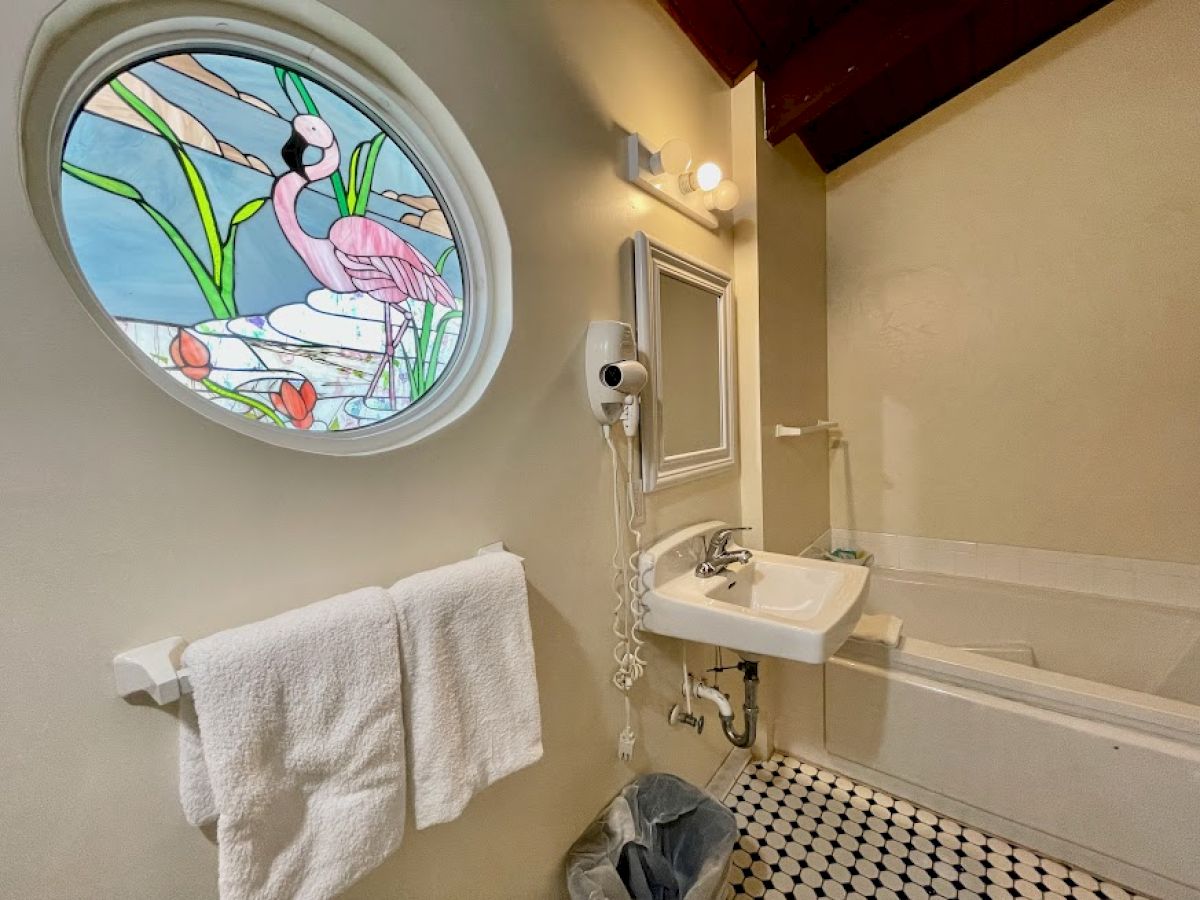 A bathroom with a sink, bathtub, towels, and a window featuring flamingo-themed stained glass. A hairdryer is mounted on the wall.