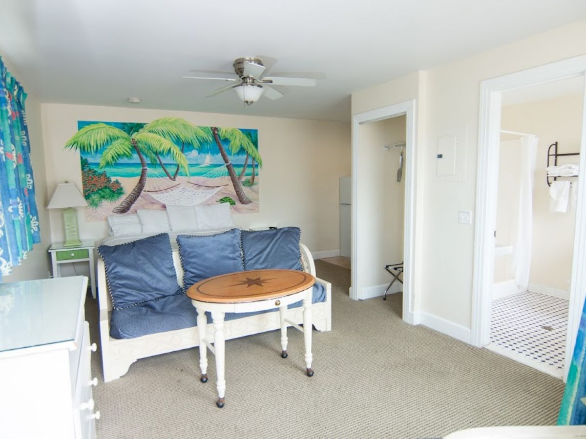 A small room with a sofa, a round coffee table, a painting of palm trees, a ceiling fan, and an adjacent clean bathroom with a checkered floor.