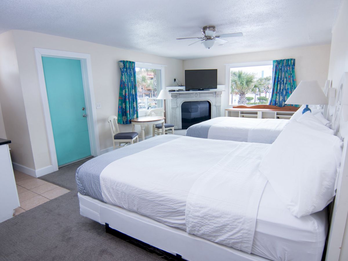 The image shows a clean, modern hotel room with two beds, a wall-mounted TV, a small table with chairs, a ceiling fan, and a window with colorful curtains.