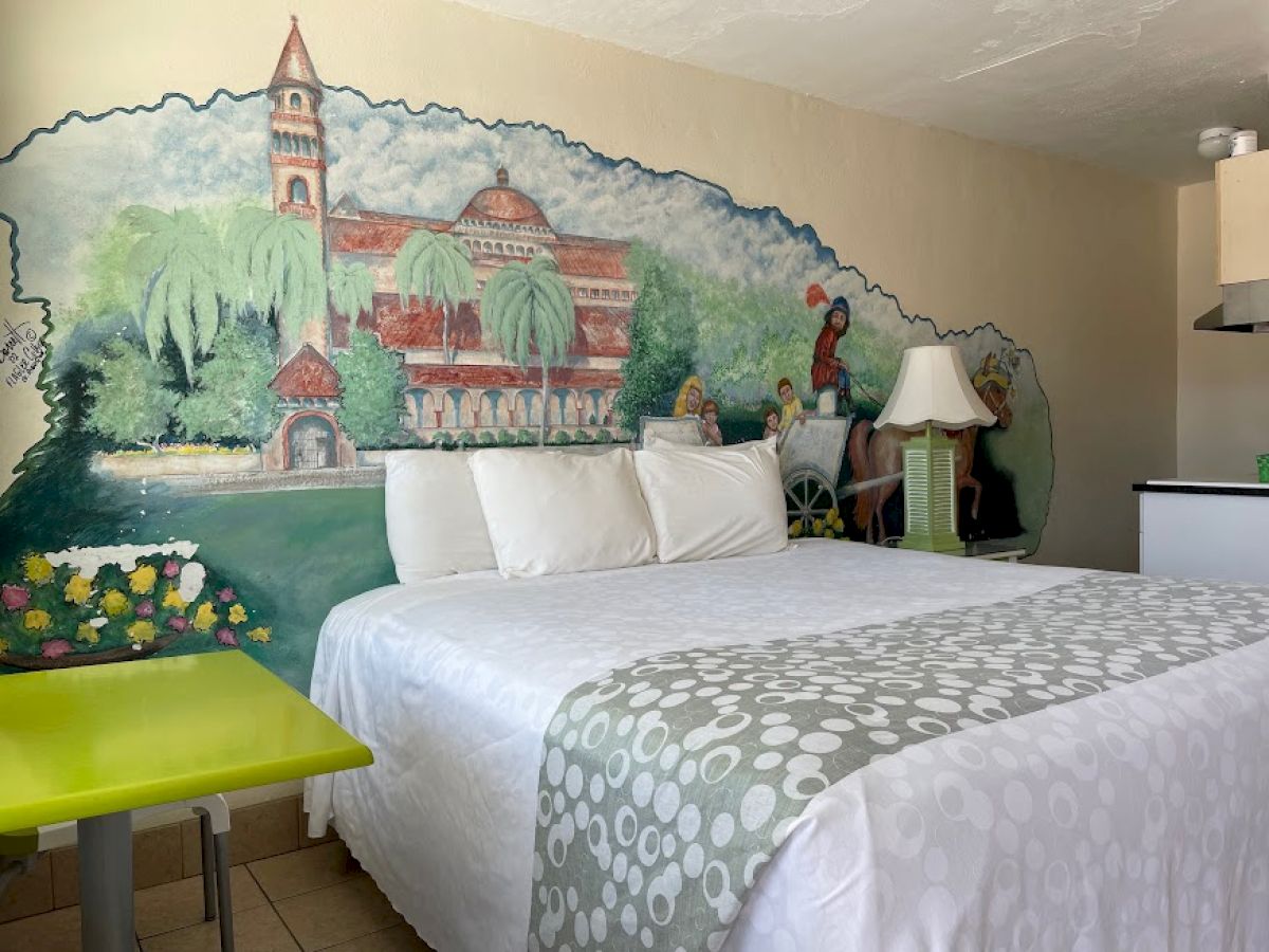 A cozy bedroom with a bed, white linens, a green table, and a wall mural depicting a picturesque building and landscape scene.