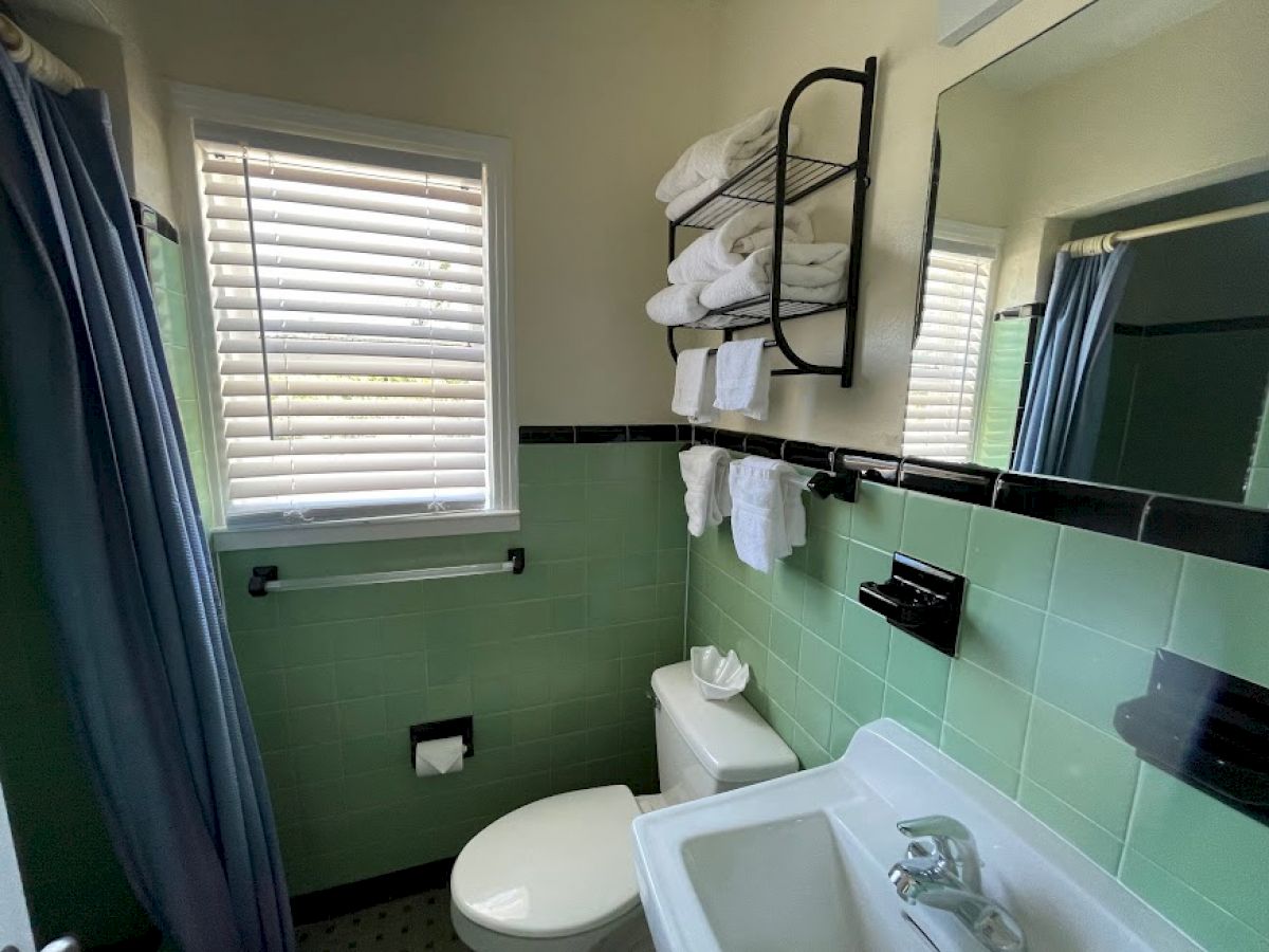 A small bathroom with a window, green tiles, a shower with a blue curtain, a toilet, a sink, a mirror, and a towel rack with towels ends the sentence.