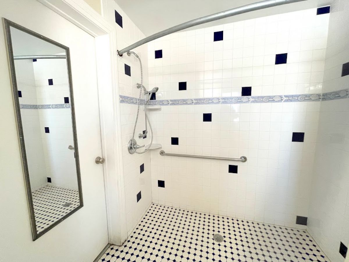 A clean, white-tiled shower with a black and white checkered floor, grab bars, a showerhead, and a mirror on the wall.