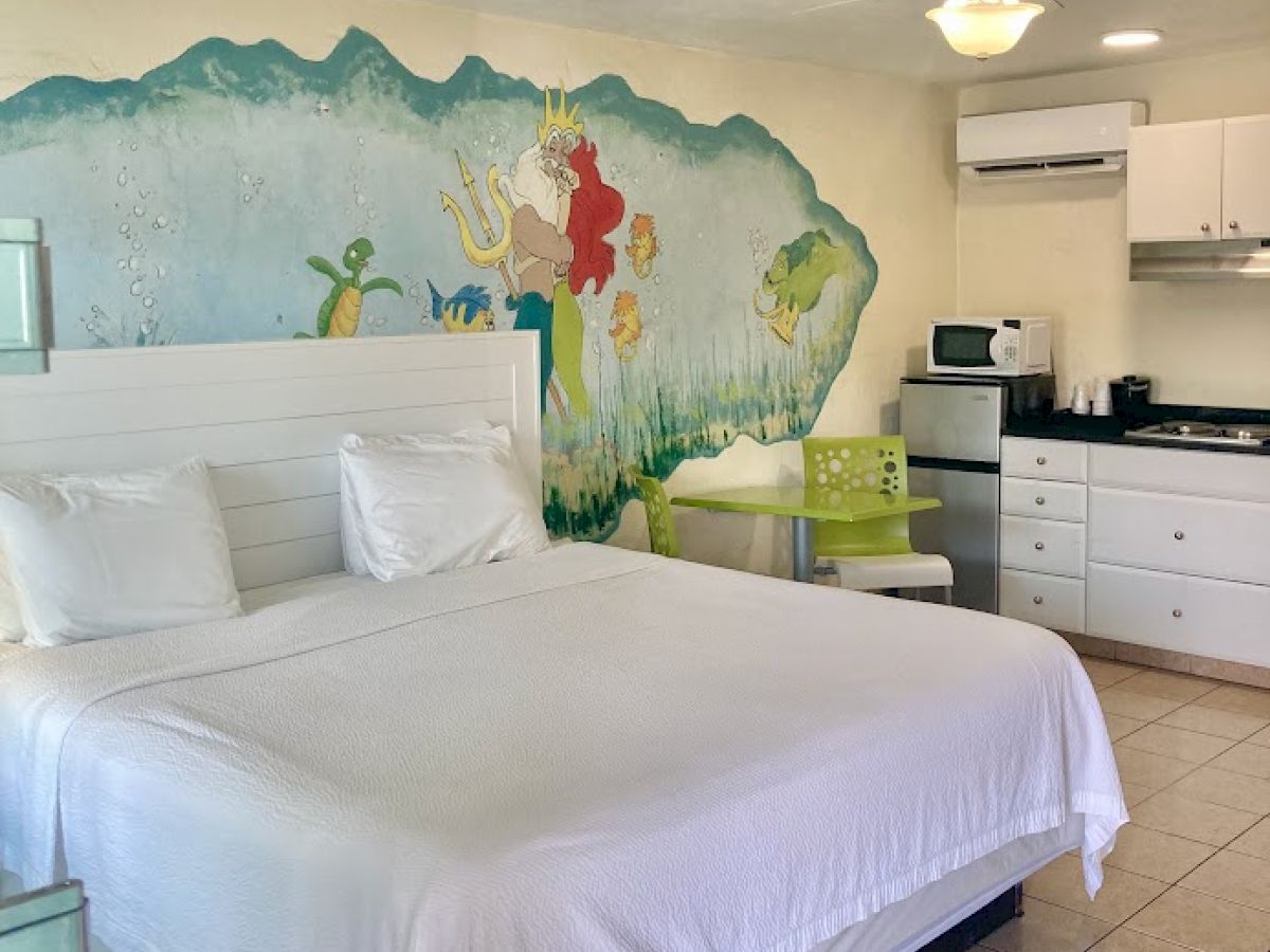 This image shows a hotel room with a bed, wall art, a small kitchenette, and a ceiling fan. The wall art features colorful sea creatures.