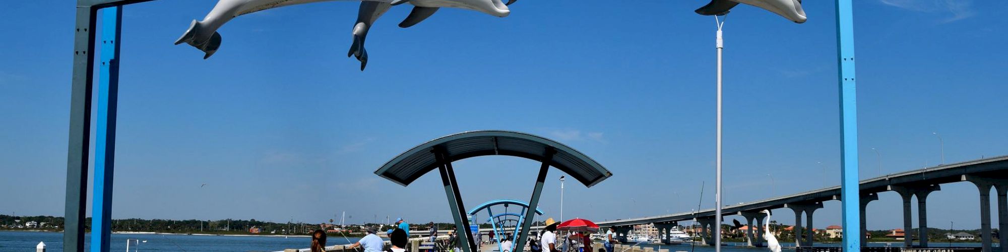 An outdoor walkway over water features a blue arch with three dolphin statues, several people, and distant buildings under a clear sky.